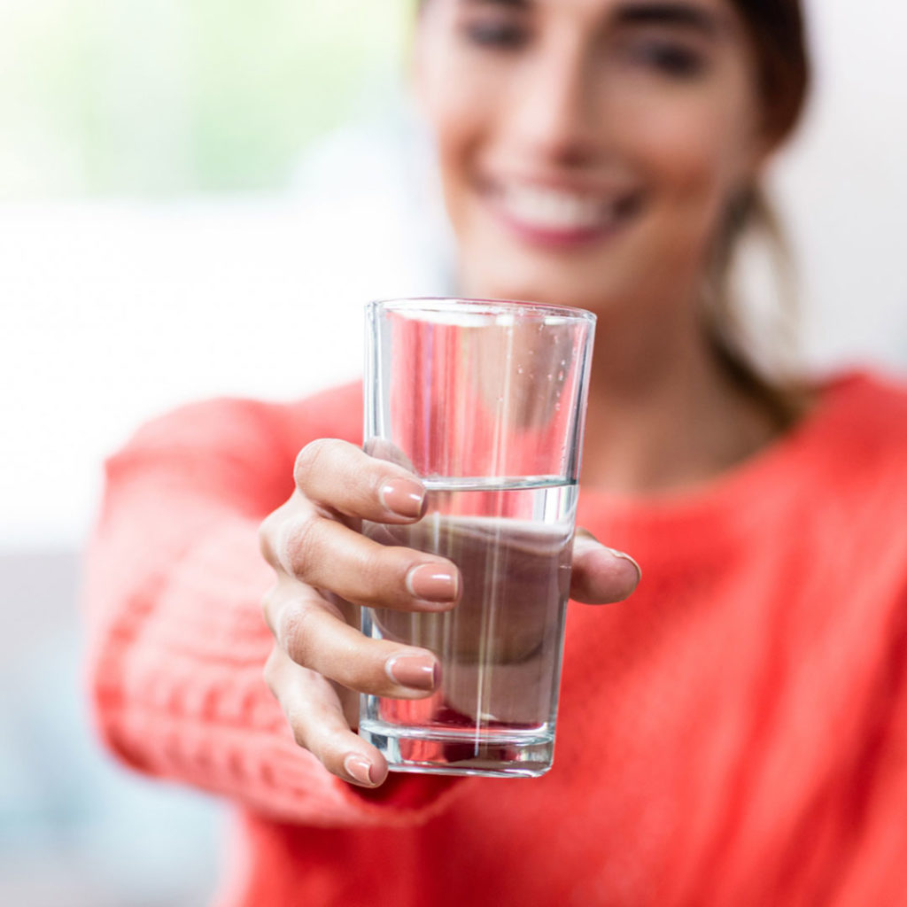 Young woman holding a glass of drinking water toward the camera.